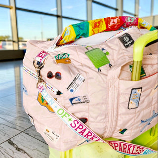 Queen of Sparkles beige travel duffle bag with sequin travel patches and rhinestone zipper attached to rolling suitcase airport view