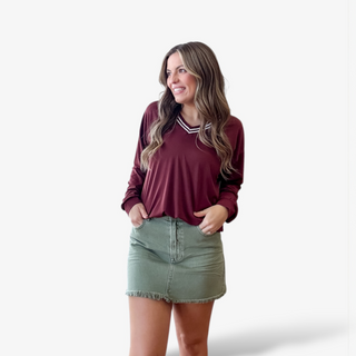 Maroon long sleeve V-neck top with striped trim styled with an olive denim skirt on a woman against a white background.