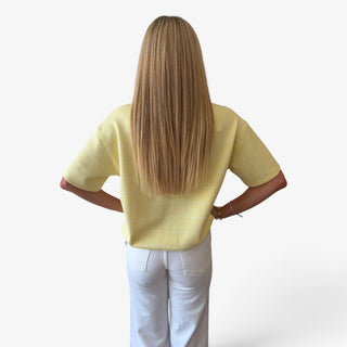 Model viewed from behind wearing a pale yellow short-sleeve scuba top with three quarter length sleeves and white pants.