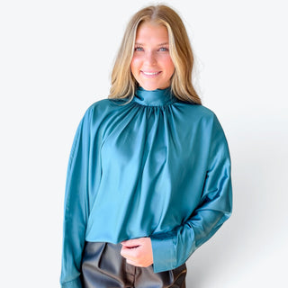 Teal satin tie back blouse with a high gathered neckline and long sleeves, styled with black faux leather shorts, front close-up view.