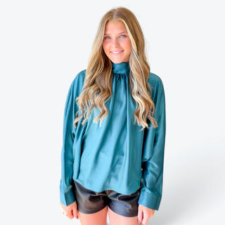 Women's teal satin long sleeve blouse featuring a high neckline, relaxed fit and smooth satin finish, styled with black shorts, full front view.