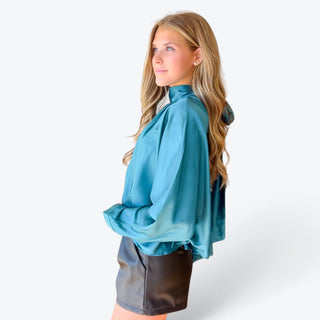 Teal satin blouse with long sleeves, wide cuffs and a relaxed draped silhouette, paired with black faux leather shorts, side profile view.