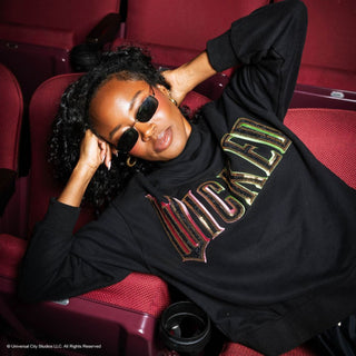 Model reclining in theater seat wearing Queen of Sparkles Licensed Black "Wicked" Logo Sweatshirt