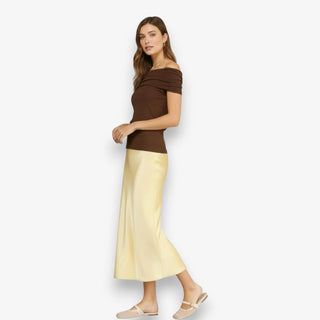 Woman wearing a butter yellow satin midi slip skirt styled with a brown top and neutral flats on a white background