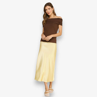 Woman wearing a butter yellow satin midi slip skirt styled with a brown top and neutral flats on a white background